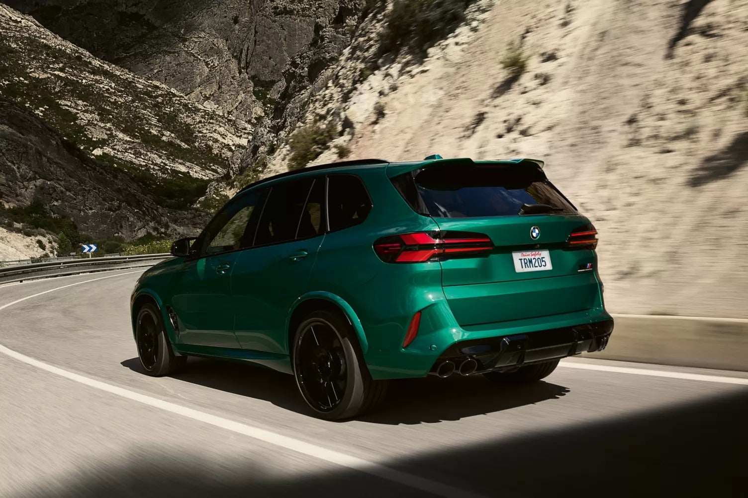 a rear view of a forest green BMW X5 driving around a hill on a curve